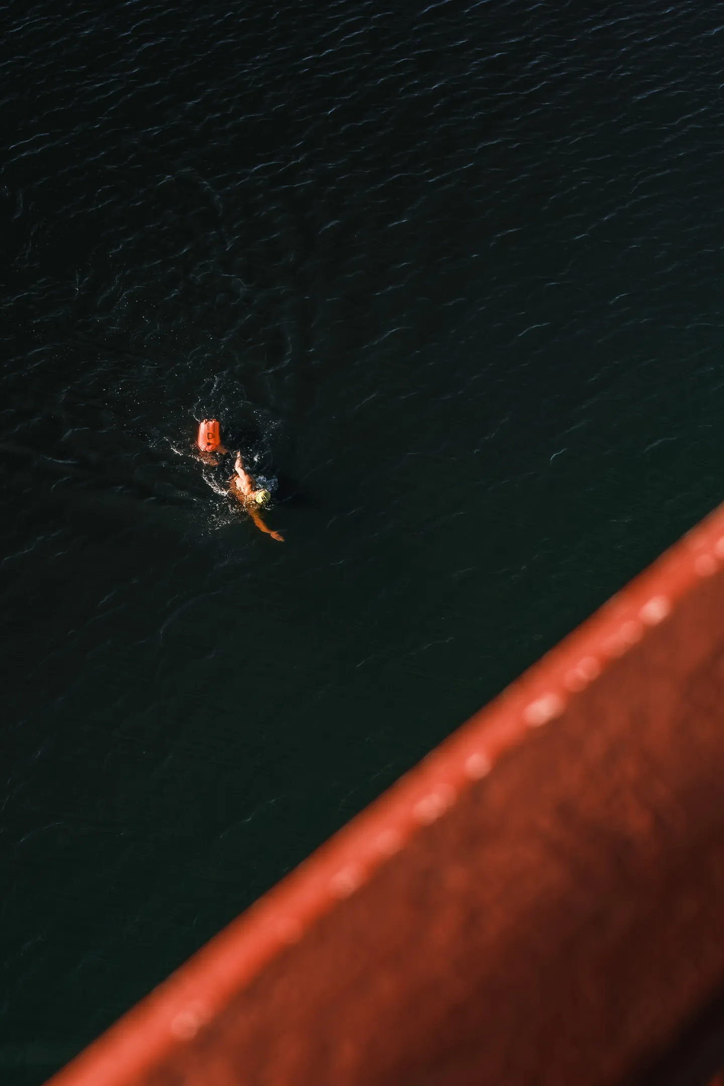 A swimmer with an orange buoy passes below the red railing.
