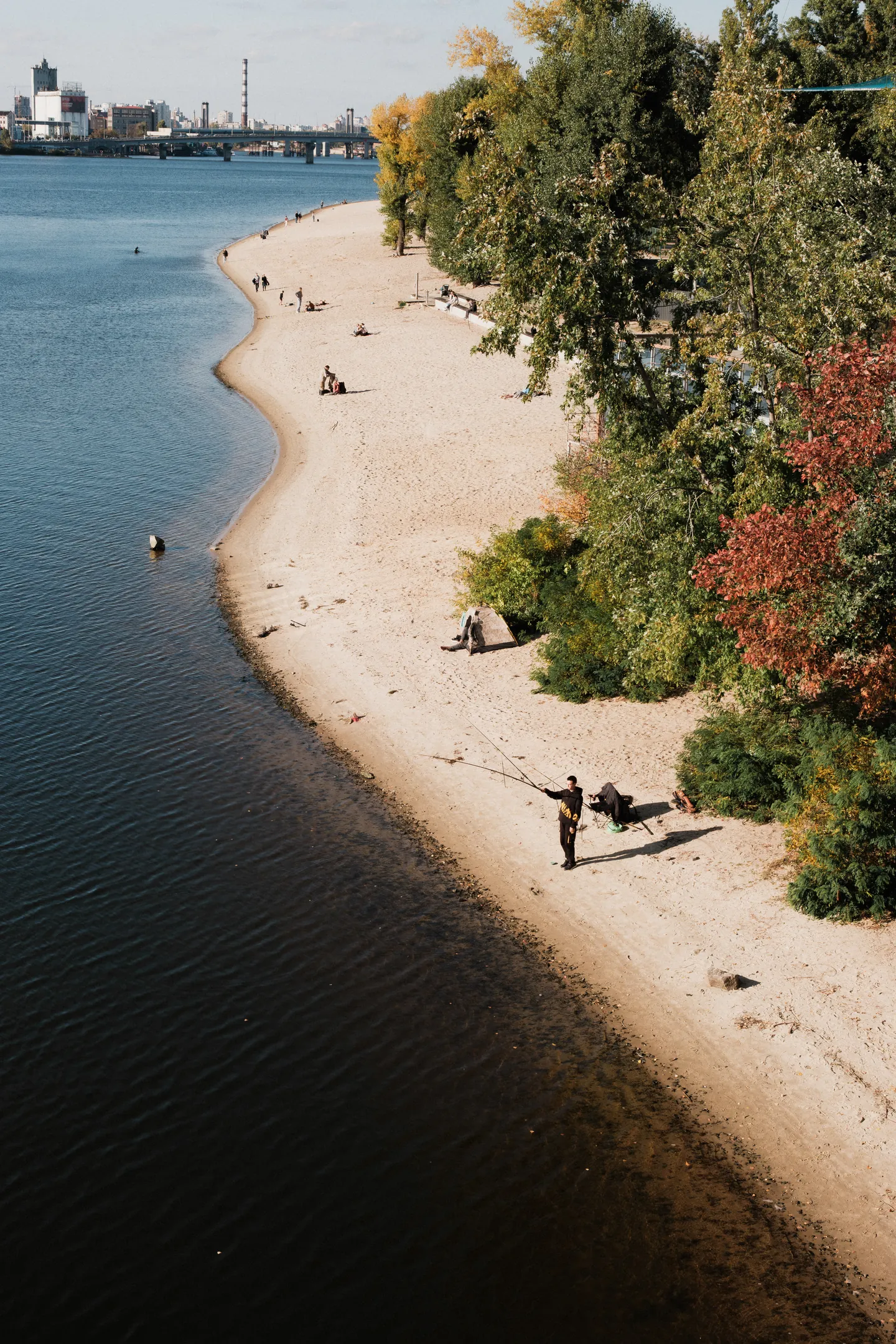 People fish, walk and rest on the beach beneath autumn trees.